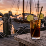 Ly rum cola với lát chanh và đá viên trên bàn gỗ bến cảng, thuyền buồm mờ phía sau lúc hoàng hôn